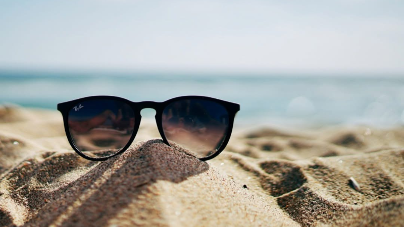 Black Ray-Ban sunglasses resting on a mound of sand at a sunny beach with the sea blurred in the background.